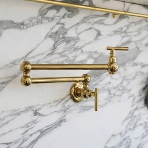 Pode incluir: Uma torneira dourada para encher panelas, montada na parede, com um design de braço oscilante. A torneira está instalada contra um backsplash de mármore. As panelas de cobre penduram de uma barra dourada acima do fogão.
