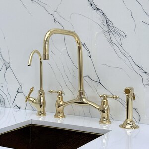 Pode incluir: Um conjunto de torneiras de cozinha douradas com um bico de arco alto, uma torneira de filtro de água separada e um pulverizador lateral. A torneira tem duas alças e está colocada contra um painel traseiro com padrão de mármore branco.