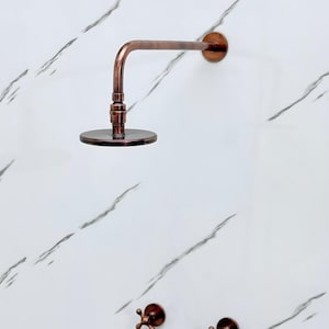 May include: A copper-colored shower head with a round, flat head and a curved arm. The shower head is mounted on a white marble-patterned wall, along with two matching faucet handles.
