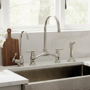 Puede incluir: Un fregadero de cocina de acero inoxidable con acabado martillado está empotrado en una encimera blanca. Un grifo y un pulverizador cromados están instalados encima del fregadero. Un jarrón blanco con flores blancas y una tabla de cortar de madera están en la encimera.