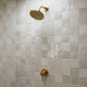 May include: A wall covered in square, glossy, off-white and beige ceramic tiles with a brass shower head and a matching brass single-handle shower control valve mounted directly onto the tiled surface in a vertical arrangement.