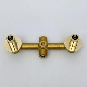 May include: A brass-coloured plumbing fixture with a horizontal bar and two circular end pieces. The fixture has a cross-shaped centre section and is designed for water control. The finish is a shiny, metallic gold.