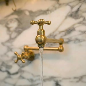 Könnte beinhalten: Nahaufnahme eines Wasserhahns aus Messing mit fließendem Wasser. Der Wasserhahn hat einen kreuzförmigen Griff und einen gebogenen Auslauf. Der Hintergrund ist eine Marmorwand.