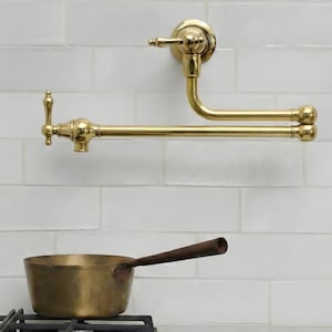Könnte beinhalten: Ein goldfarbener, an der Wand montierter Topffüller-Wasserhahn mit gebogenem Auslauf und dekorativen Griffen. Der Wasserhahn ist an einer weiß gefliesten Wand installiert. Ein Messingtopf mit Holzgriff steht auf einem Gasherd.