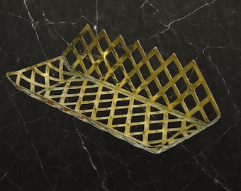 May include: A gold-colored, rectangular basket with a diamond lattice design. The basket has a zig-zag edge on one side. The basket is made of metal and is set against a black marble background.