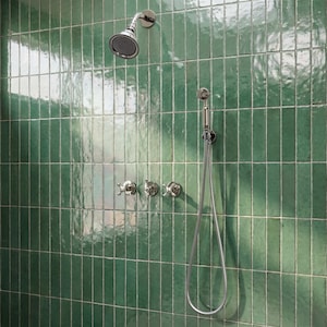 Puede incluir: Una moderna instalación de ducha con un cabezal de ducha cromado y un pulverizador de mano, sobre un fondo de baldosas rectangulares verde esmeralda brillantes. Incluye tres manijas de grifo en forma de cruz.