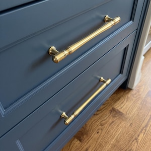 May include: Two horizontal drawer pulls made of polished brass with a cylindrical shape and subtle grooved detailing at the ends. These metallic handles are mounted onto a dark navy blue cabinet with a recessed panel design and smooth finish.