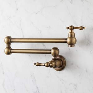Puede incluir: Un grifo de pared para llenar ollas con un brazo oscilante de doble articulación y un acabado en latón cepillado. El accesorio cuenta con dos manijas de palanca con extremos decorativos redondeados y un caño central para la cocina.
