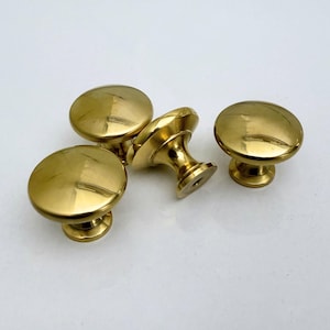 Könnte beinhalten: Vier polierte Messing-Schrankknöpfe. Jeder Knopf hat eine abgerundete Oberseite und einen kurzen, zylindrischen Sockel. Ein Knopf ist auf der Seite gedreht und zeigt den Schaft und das Schraubenloch. Goldene Farbe.