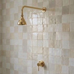 May include: A wall-mounted shower system featuring a polished brass finish, including a bell-shaped shower head on a horizontal arm and a single-handle control valve, installed against a wall of square, glossy, off-white and beige ceramic tiles.