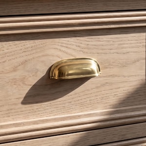 May include: A polished brass cup pull handle mounted on a light-toned wooden drawer front. The handle features a smooth, curved, semi-circular design with a metallic finish that reflects light, contrasting against the natural grain of the wood.