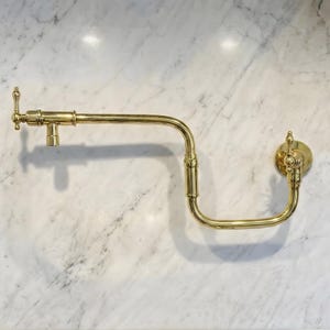 May include: A gold-colored, wall-mounted pot filler faucet with a curved, articulated arm. The faucet features a decorative handle and a round wall plate. The fixture is made of brass and is installed on a white marble surface.