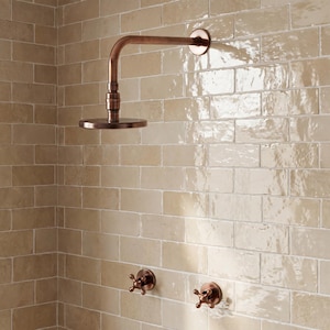 Op de afbeelding: Een koperkleurige douchekop en kraanarmaturen zijn gemonteerd op een beige betegelde muur. De douchekop heeft een cirkelvormig ontwerp en is verbonden met een gebogen pijp. Twee kruisvormige kraanhendels bevinden zich eronder.