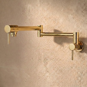Pode incluir: Uma torneira dourada para encher panelas, montada na parede. A torneira tem um braço longo e articulado que se estende da parede. Possui um corpo cilíndrico, um bico e uma alavanca. O acabamento é dourado brilhante e polido.