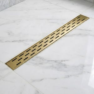 May include: A rectangular, gold-colored shower drain with a series of evenly spaced, oblong openings. The drain is set into a white marble tile floor, providing a sleek and modern aesthetic for bathroom fixtures.