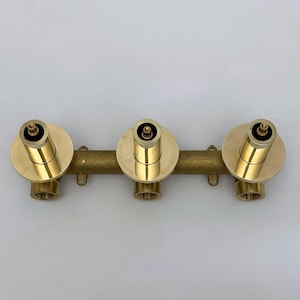 May include: A brass shower valve with three circular faucet heads. Each head has a cylindrical design with a circular base. The valve is connected by a horizontal pipe with mounting loops.