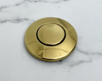 May include: A close-up of a gold-colored, circular button with a raised center. The button has a shiny, metallic finish and is set against a white and gray marble background. The button is approximately 1.5 inches in diameter.