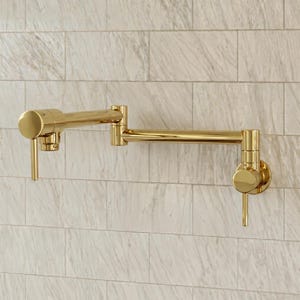 Puede incluir: Un grifo de llenado de ollas montado en la pared con un acabado en oro pulido. El accesorio presenta un diseño de brazo oscilante articulado de doble articulación con dos manijas cilíndricas, una en la base y otra en la salida para el control.