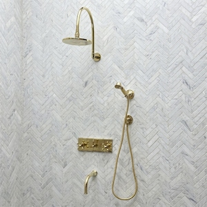 May include: A gold-colored shower fixture set against a white and gray herringbone tile wall. The set includes a rain shower head, a handheld shower, and a faucet. The fixtures have a classic, elegant design.