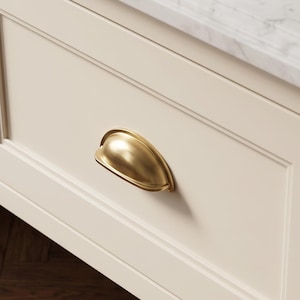 May include: A brushed brass cup pull handle mounted on a cream-colored cabinet drawer. The hardware features a smooth, semi-circular design with a metallic finish, attached to a flat-front drawer panel with recessed trim details and a marble top.