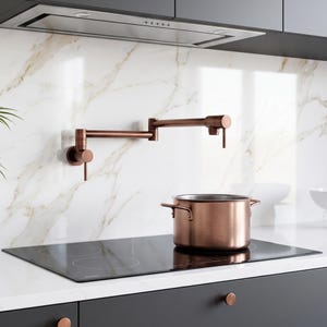 May include: A wall-mounted copper pot filler faucet with a dual-jointed swing arm and a matching copper stockpot sitting on a black glass induction cooktop. The kitchen features a white marble backsplash with grey veining and dark cabinetry.