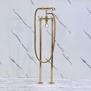 May include: A gold-coloured freestanding bath tap with a handheld showerhead. The tap has cross-shaped handles and a flexible hose. The fixture is set against a white marble-patterned background.