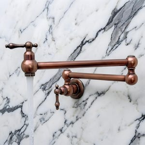 May include: A copper-colored, wall-mounted pot filler faucet with a two-jointed, articulated arm. Water flows from the faucet against a white and gray marble background. The faucet has a vintage design with decorative handles.