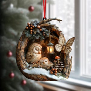 Könnte beinhalten: Ein hängendes Ornament in Form eines hohlen Baumstamms mit zwei braunen Eulen mit geschlossenen Augen, einer kleinen leuchtenden Laterne, einem großen beigen Schmetterling, Tannenzapfen, Beeren und weißem Schnee, befestigt an einem roten Band.