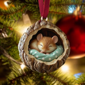 May include: A Christmas ornament featuring a sleeping mouse nestled in a walnut shell. The mouse rests on a light blue pillow with white polka dots. The ornament is suspended by a maroon ribbon, with a blurred Christmas tree background.