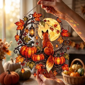 May include: A stained glass suncatcher featuring a fox sitting before a full moon, surrounded by orange pumpkins, autumn maple leaves, and a small hanging lantern. The circular design is crafted with intricate metal framing and colourful glass.