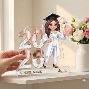 Puede incluir: Figura acrílica de graduación con una enfermera de dibujos animados con bata y gorro blancos, sosteniendo un diploma. Los números "2026" son en oro rosa y mármol. La base dice "SCHOOL NAME" y "Julia".