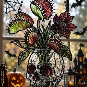 May include: A stained glass suncatcher featuring a clear, textured skull-shaped vase holding several green and red Venus flytrap plants and a dark red flower. The piece is framed in black metal and hangs from a chain, displaying vibrant colors.