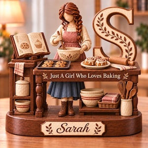 Può includere: Una statuetta decorativa di una persona che cucina, con una grande lettera S, un ricettario, un vassoio di biscotti e un tavolo con il testo Just A Girl Who Loves Baking. La base riporta il nome Sarah, con vari utensili da cucina e ciotole.