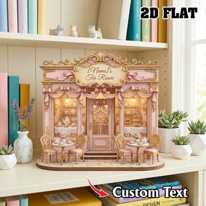 Könnte beinhalten: Ein rosa-goldenes 2D-Flachmodell eines Teezimmers mit dem Text "[Name]'s Tea Room". Das Modell hat eine detaillierte Fassade mit Tischen, Stühlen und dekorativen Elementen. Es wird auf einem Regal mit Büchern und Pflanzen ausgestellt.