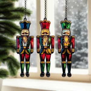 May include: Three hanging nutcracker ornaments featuring stained glass style panels in red, green, blue, and yellow. Each figure has a black metal frame, a moustache, and a gold-toned belt, suspended by a black metal chain for holiday decoration.
