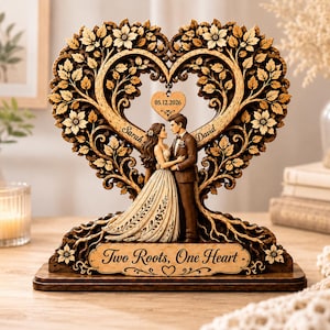 Può includere: Una scultura decorativa in legno con una sposa e uno sposo in piedi insieme. Le figure sono incorniciate da un albero a forma di cuore con foglie e fiori. Un cuore sospeso mostra 05.12.2026, i nomi Sarah e David, e il testo Two Roots, One Heart.