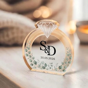 May include: A clear acrylic ring-shaped ornament with a diamond-shaped top. The ring features the initials "S&D" and the date "20.05.2026" in black, surrounded by a floral design with green leaves. A wedding or engagement themed item.