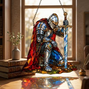 May include: A stained glass suncatcher depicting a knight in silver armor kneeling and holding a long sword. The knight wears a vibrant red cape and blue accents. The piece features intricate lead lines and colorful glass segments, suspended by a metal chain.