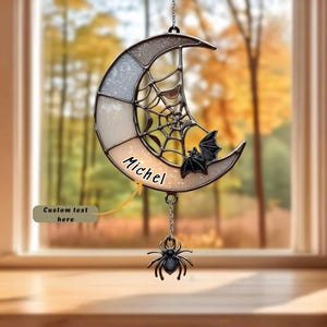 May include: A crescent moon-shaped stained glass suncatcher featuring a spiderweb pattern, a small black bat, and a dangling spider charm. The piece includes the name Michel printed in black text on the lower curve of the moon frame.