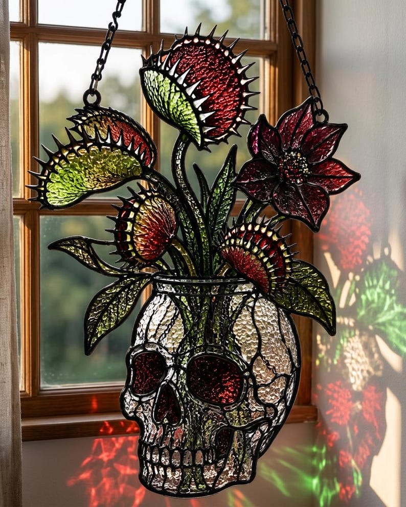 May include: A stained glass suncatcher featuring a clear textured skull-shaped vase holding several red and green Venus flytrap plants and a dark red flower. The piece is framed in black metal and hangs from a black chain against a window.