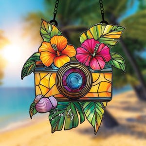 Peut inclure: Décoration en vitrail représentant un appareil photo orné de fleurs d'hibiscus orange et rose, de feuilles vertes et d'un coquillage. L'objectif de l'appareil photo est bleu et violet. Le nom "Emily" est écrit sur une feuille.