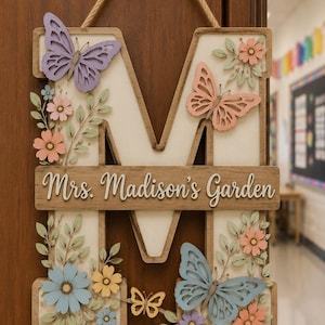 May include: A wooden wall hanging featuring a large letter M with a cream-coloured background. The letter is decorated with layered cutouts of butterflies in purple, peach, and blue, along with small flowers and leaves. A horizontal wooden plaque across the centre displays the text Mrs. Madison's Garden in white script. A jute rope is attached to the top for hanging.