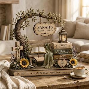May include: A decorative tabletop display featuring a sculpted olive tree with a hanging sign reading SARAH'S PRAYER GARDEN, a bench with an open book, a cross, sunflowers, a lantern, and stacked books labelled TRUST, STRENGTH, and LOVE.