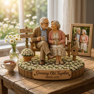 Op de afbeelding: Een decoratief beeldje van een ouder echtpaar dat op een bankje zit met mokken in hun handen. De basis bevat de tekst "Growing Old Together SINCE 1972" en een bordje met "Our Story" en "Our Forever". Inclusief een vogelhuisje en madeliefjes.