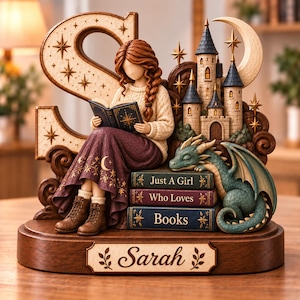 May include: A decorative figurine featuring a person with braided hair reading a book, seated on a stack of three books labelled Just A Girl, Who Loves, Books. A green dragon rests beside them, with a castle, a crescent moon, and the letter S behind.