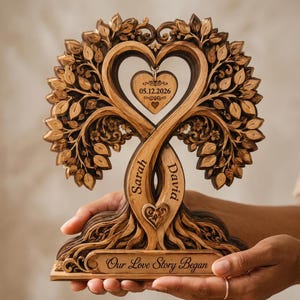 May include: A carved wooden tree sculpture featuring two intertwined trunks engraved with the names Sarah and David. A heart-shaped pendant hangs from the branches with the date 05.12.2026, and the base is inscribed with the text Our Love Story Began.