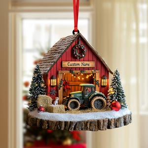 Puede incluir: Un adorno navideño colgante con forma de granero rojo y un tractor verde en su interior. El granero tiene una corona, luces y un letrero que dice Custom Name. Se asienta sobre una base de madera con nieve, fardos de heno y dos pinos.