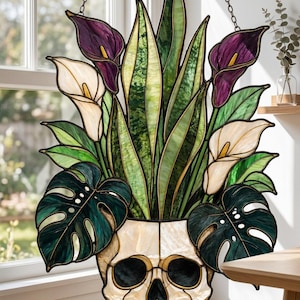May include: A stained glass suncatcher featuring a white skull base holding a vibrant arrangement of green snake plant leaves, dark green monstera leaves, and purple and white calla lily flowers, all connected by dark metal solder lines.