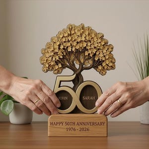 May include: A wooden 50th anniversary tree sculpture. The tree is brown with gold accents, and the number 50 is in gold with the names "John" and "Sarah". The base reads "Happy 50th Anniversary 1976-2026".