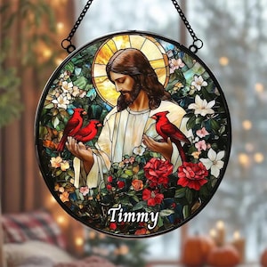 May include: A circular stained glass suncatcher featuring a depiction of a bearded figure in white robes with a golden halo. Two red cardinal birds rest on the figure's hands, and one sits on the shoulder. The name Timmy is printed at the bottom.